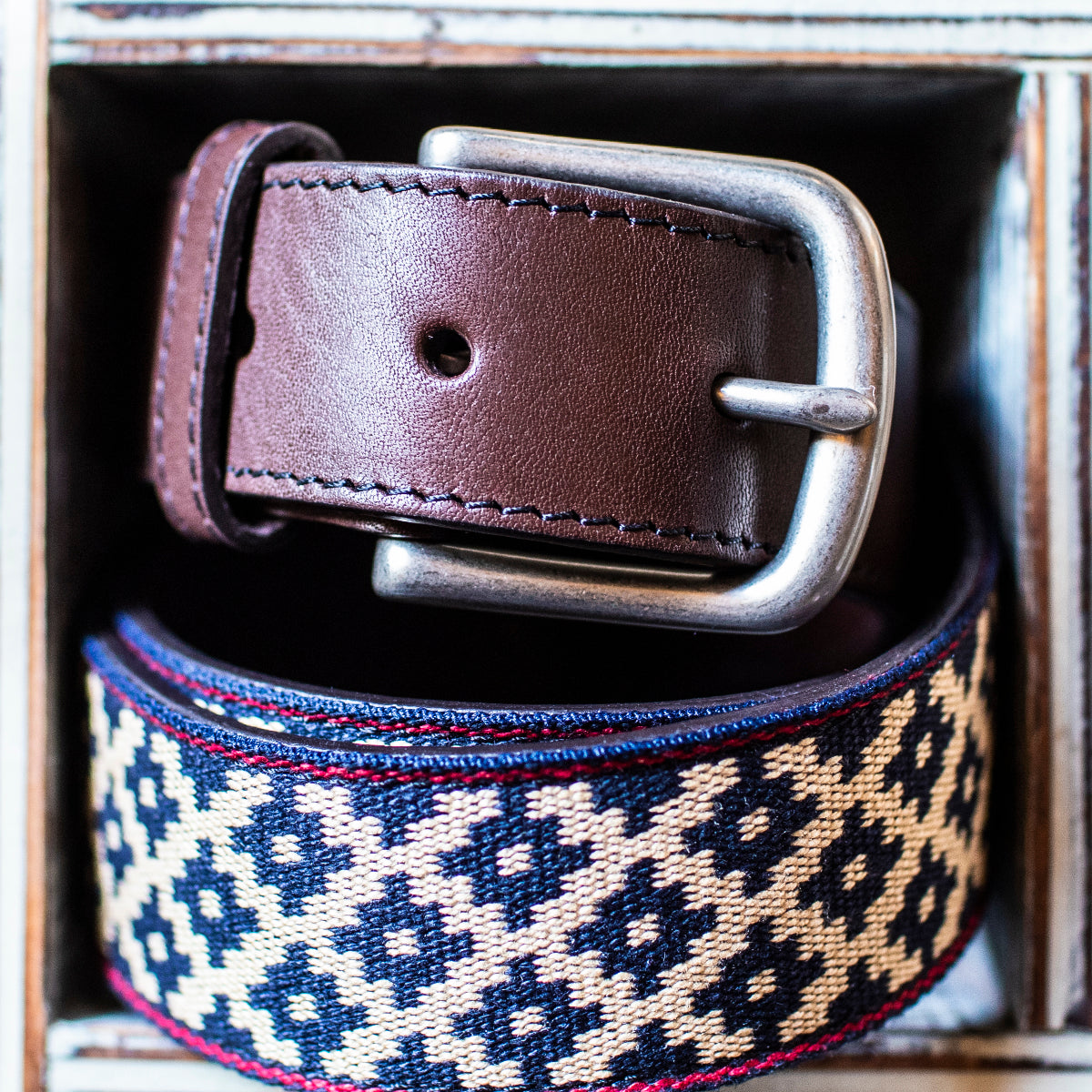 A dark brown leather belt featuring a navy blue and beige geometric woven pattern with red stitching.